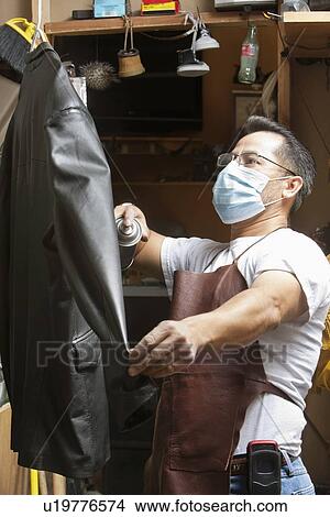 Picture - Mature cobbler wearing mask looking at leather jacket in workshop. Fotosearch