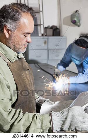 Mature man reading paper while young male welding in background View Large Photo Image Picture - Mature man reading paper while young male welding in background. Fotosearch