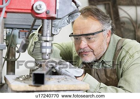 Stock Image - Mature man wearing protective eyewear while working with machinery in workshop. Fotosearch