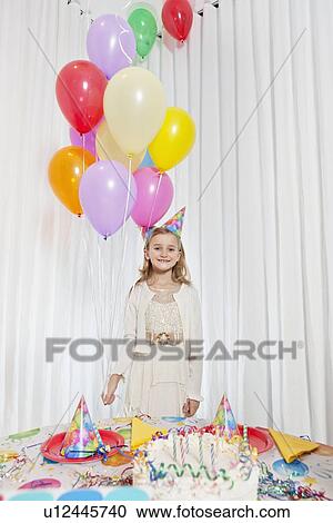 Stock Image - Portrait of a happy girl holding party balloons with cake on table. Fotosearch