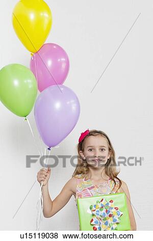 Stock Photo - Portrait of a happy girl with party balloons and gift over colored background. Fotosearch