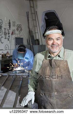 Portrait of a happy mature man wearing protective mask with young male welding in background View Large Photo Image Stock Photo - Portrait of a happy mature man wearing protective mask with young male welding in background. Fotosearch