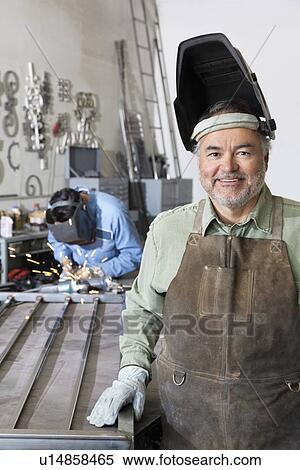 Stock Photography - Portrait of a happy mature man wearing welding mask with young male working in background. Fotosearch