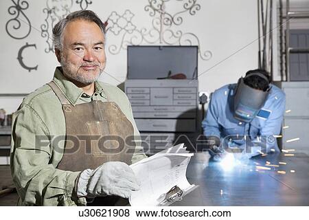 Stock Photo - Portrait of a happy mature man with young male welding in background. Fotosearch