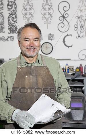 Stock Image - Portrait of a happy mature metalworker with clipboard in workshop. Fotosearch