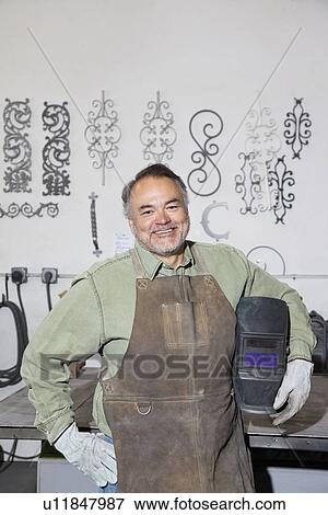 Stock Photo - Portrait of a happy mature metalworker holding protective mask in workshop. Fotosearch