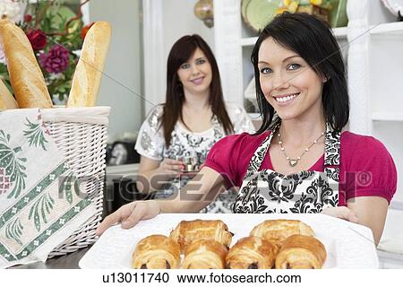 Stock Image - Portrait of a happy mid adult caterer holding pastry tray with coworker in background. Fotosearch