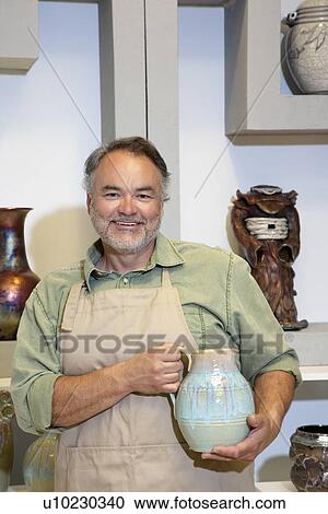 Stock Image - Portrait of a happy potter holding ceramic pot in artist's studio. Fotosearch