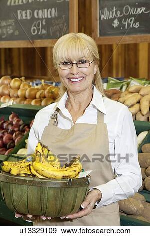Stock Photo - Portrait of a happy senior female employee carrying bananas in farmer's market. Fotosearch