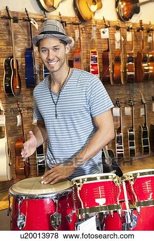 Portrait of a happy young man playing bongo drums with guitars on display in background View Large Photo Image Stock Photo - Portrait of a happy young man playing bongo drums with guitars on display in background. Fotosearch