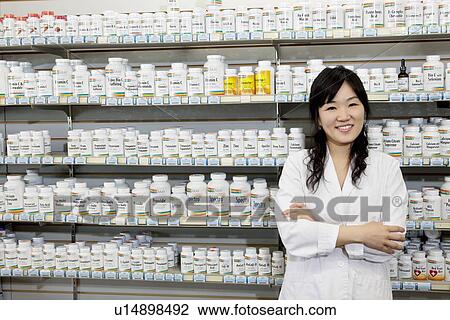 Stock Image - Portrait of a happy young woman with arms crossed in front of pills shelf in drugstore. Fotosearch