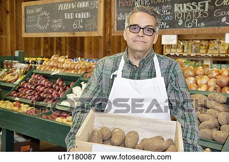 Stock Photo - Portrait of a mature employee carrying potato box in front of vegetable stand in market. Fotosearch