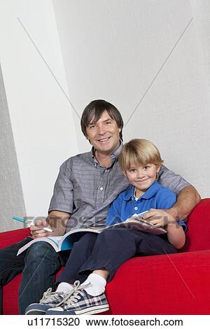 Stock Image - Portrait of father and son doing homework together while sitting on sofa. Fotosearch