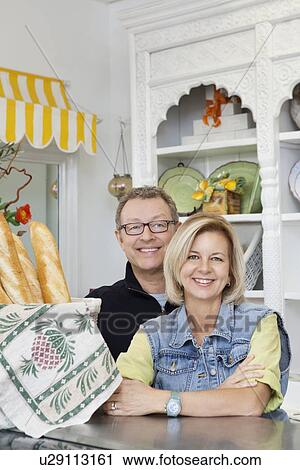 Stock Image - Portrait of happy mature couple standing at counter in cake shop. Fotosearch