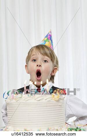 Stock Photography - Portrait of young boy wearing party hat blowing candles on birthday cake. Fotosearch