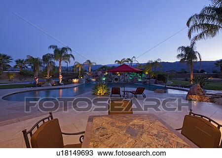 Stock Image - Swimming pool with chaise lounge outside mansion at dusk. Fotosearch