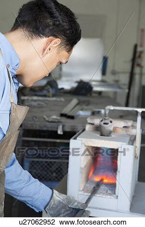 Stock Image - Young blacksmith burning metal rod in workshop. Fotosearch