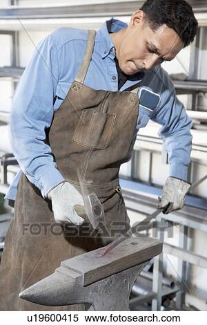 Stock Photography - Young blacksmith hammering metal rod on anvil in workshop. Fotosearch