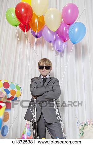 Stock Image - Young boy wearing sunglasses and standing with arms crossed holding party balloons. Fotosearch