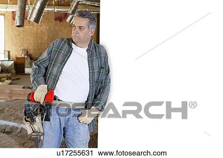 Stock Image - Construction worker holding drill while leaning on white wall. Fotosearch