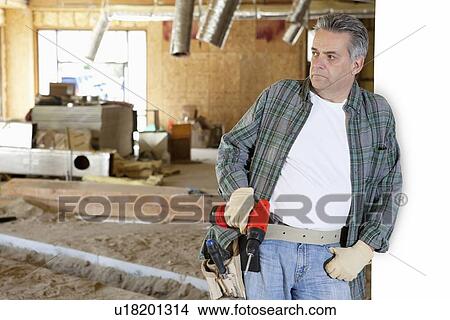 Picture - Front view of construction worker holding drill while leaning on white wall. Fotosearch