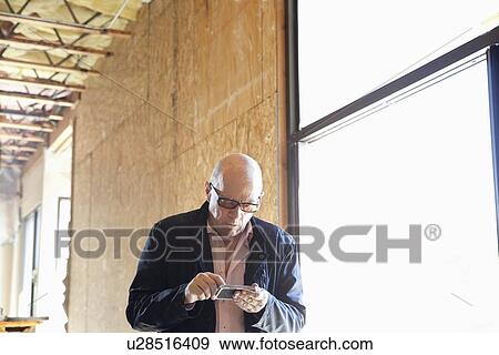 Stock Photo - Front view of senior property owner text messaging near window. Fotosearch