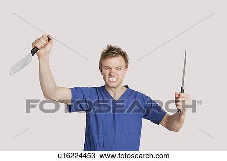Stock Image - Portrait of angry male nurse with knives in both hands. Fotosearch