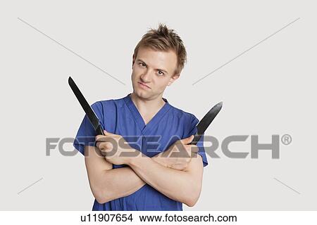 Portrait of angry young male nurse with knives standing over light gray background View Large Photo Image Picture - Portrait of angry young male nurse with knives standing over light gray background. Fotosearch