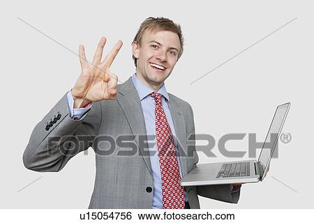 Stock Photograph - Portrait of cheerful businessman with laptop gesturing ok sign over colored background. Fotosearch