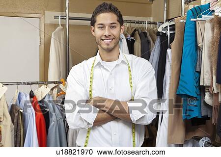 Stock Image - Portrait of handsome young tailor with arms crossed standing in front of clothes rail. Fotosearch