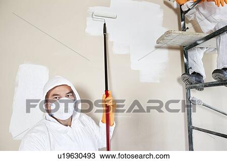 Stock Image - Portrait of male worker holding paint roller with female worker sitting on scaffold. Fotosearch