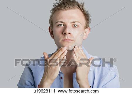 Stock Image - Portrait of young man applying lotion on face over colored background. Fotosearch