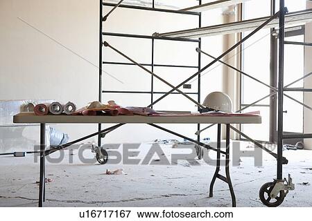 Stock Photo - Table with blueprints and hardhat in front of scaffold in construction room. Fotosearch