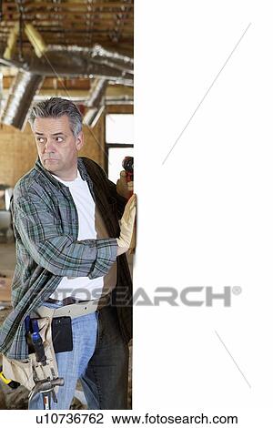 Stock Image - Worker holding drill standing next to white wall. Fotosearch