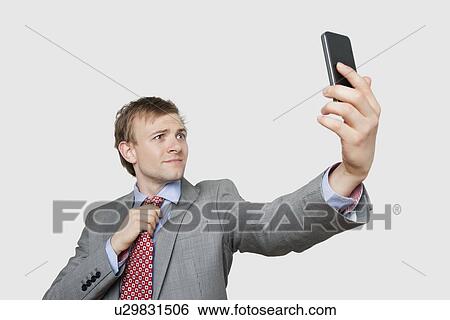 Stock Photograph - Young businessman taking self photograph while adjusting tie over colored background. Fotosearch