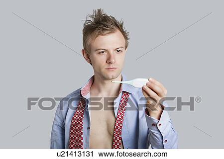 Stock Image - Young man looking at thermometer over colored background. Fotosearch