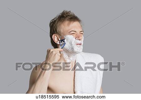 Young man shaving while making faces with eyes closed over colored background View Large Photo Image Stock Photography - Young man shaving while making faces with eyes closed over colored background. Fotosearch