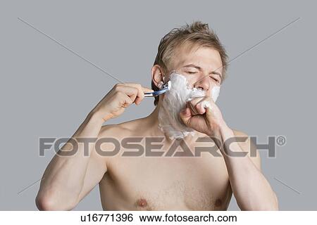 Stock Photograph - Young shirtless man covering mouth while shaving over colored background. Fotosearch