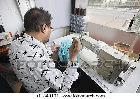 Stock Image - Back view of male tailor stitching cloth while sitting at sewing machine. Fotosearch