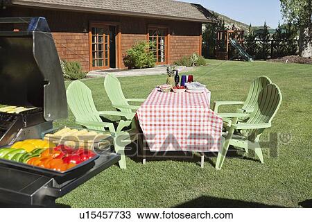 Stock Image - Dining table and chairs with barbecue grill on lawn in front of country house. Fotosearch