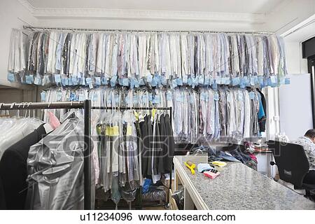 Stock Photograph - Dry cleaned shirts hanging on clothes rack while tailor working in corner of the room. Fotosearch
