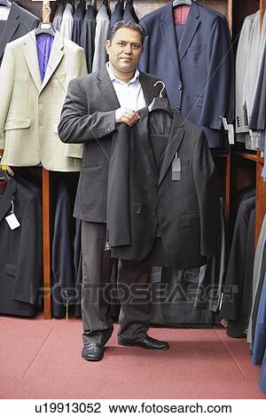 Stock Image - Full length portrait of man shopping for formal coat in menswear store. Fotosearch