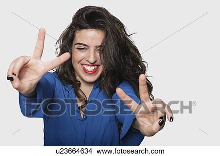 Picture - Happy young woman gesturing victory sign against gray background. Fotosearch