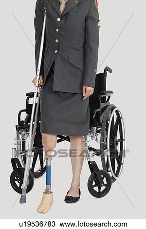 Stock Image - Low section of a female US military officer with crutch standing in front of wheelchair over gray background. Fotosearch