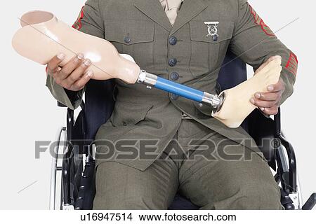 Picture - Midsection of a US military officer holding prosthesis leg as he sits in wheelchair. Fotosearch