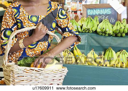 Stock Image - Midsection of African American in traditional wear with vegetable basket at super market. Fotosearch