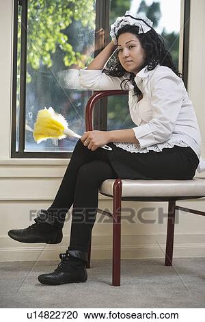 Stock Image - Pensive housemaid sitting on chair with a feather duster. Fotosearch