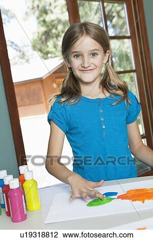 Stock Image - Portrait of a little girl finger painting on paper at table. Fotosearch