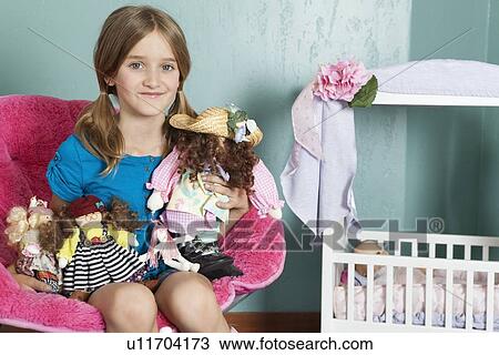 Stock Image - Portrait of a little girl with stuffed toys sitting on chair by doll's crib. Fotosearch