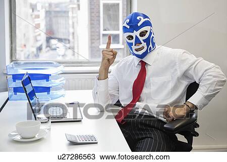 Stock Image - Portrait of businessman in wrestling mask points up at office desk. Fotosearch